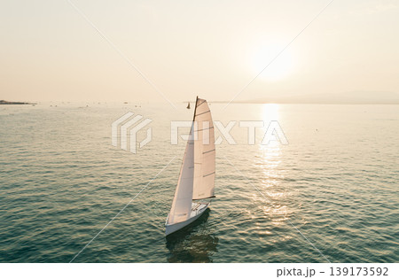 Aerial view captures solitary white sailboat gracefully navigates tranquil waters reflecting the warm glow of a beautiful sunset Aerial view captures solitary white sailboat gracefully navigates tranquil waters reflecting the warm glow of a beautiful sunset 139173592