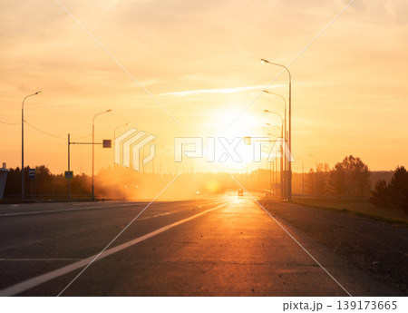 Golden hour light bathes a long, straight road lined with streetlights, leading to a bright horizon and a distant car at sunset Golden hour light bathes a long, straight road lined with streetlights, leading to a bright horizon and a distant car at sunset 139173665