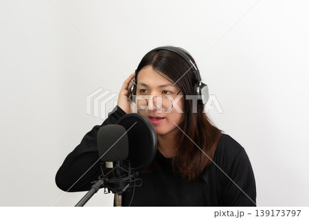 Confident LGBTQ Asian woman wearing headphones speaking into microphone in studio minimal background modern content creator lifestyle focused calm expression Confident LGBTQ Asian woman wearing headphones speaking into microphone in studio minimal background modern content creator lifestyle focused calm expression 139173797