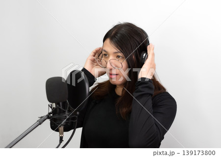 Confident LGBTQ Asian woman wearing glasses adjusting headphones while recording voice with microphone in studio minimal background modern content creator lifestyle focused expression 139173800