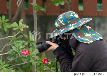 Photographer capturing red flowers with camera in garden wearing hat and protective clothing showing outdoor hobby nature exploration creativity and focus 139173817