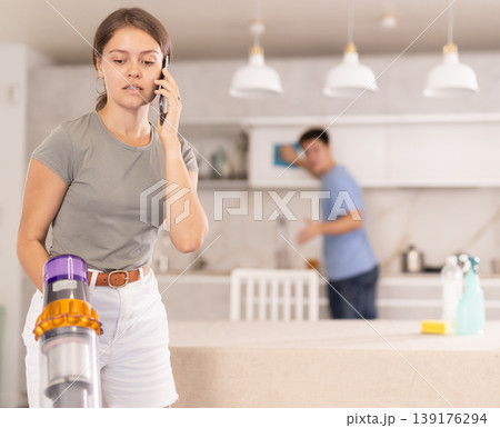 Girl cleans kitchen with vacuum cleaner and has nice conversation on phone with her friend Girl cleans kitchen with vacuum cleaner and has nice conversation on phone with her friend 139176294