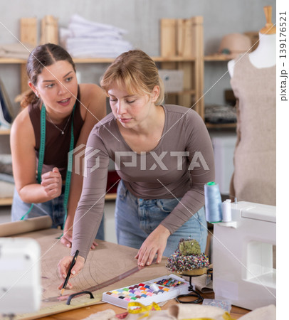 Two young female dressmakers drawing on paper in sewing workshop Two young female dressmakers drawing on paper in sewing workshop 139176521