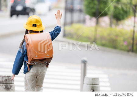 ランドセルを背負って道路の横断歩道を手をあげて渡る小学生の後ろ姿、登校や通学の交通安全イメージ顔無し 139176876