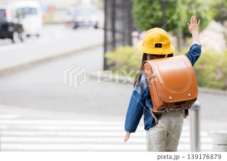 ランドセルを背負って道路の横断歩道を手をあげて渡る小学生の後ろ姿、登校や通学の交通安全イメージ顔無し 139176879