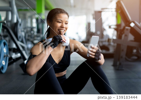 Black woman sitting on gym floor eating snack bar and using smartphone Black woman sitting on gym floor eating snack bar and using smartphone 139183444