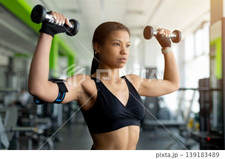 Focused young Black woman doing overhead dumbbell shoulder press in gym Focused young Black woman doing overhead dumbbell shoulder press in gym 139183489