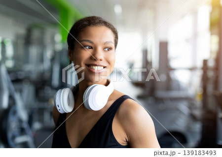 Smiling young Black woman in gym with white wireless headphones around neck Smiling young Black woman in gym with white wireless headphones around neck 139183493