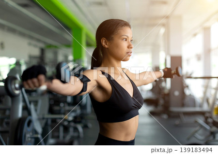 Focused young Black woman doing lateral dumbbell raises in gym Focused young Black woman doing lateral dumbbell raises in gym 139183494