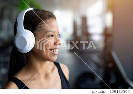 Portrait of smiling young Black woman in gym wearing white wireless headphones Portrait of smiling young Black woman in gym wearing white wireless headphones 139183500