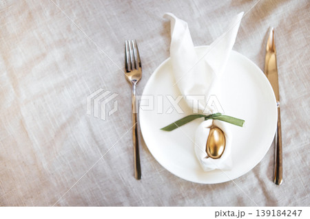 Elegant table setting featuring a white plate with a golden egg wrapped in a napkin tied with green ribbon, accompanied by a fork and knife on a textured linen tablecloth Elegant table setting featuring a white plate with a golden egg wrapped in a napkin tied with green ribbon, accompanied by a fork and knife on a textured linen tablecloth 139184247