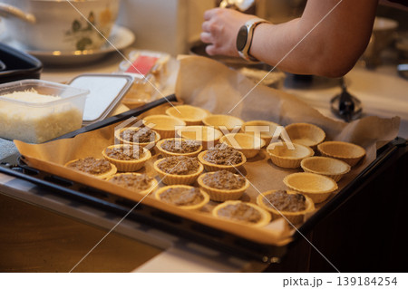 Baking process with a close-up of a tray filled with unbaked tart shells and a hand adding filling in a cozy kitchen setting with various utensils visible Baking process with a close-up of a tray filled with unbaked tart shells and a hand adding filling in a cozy kitchen setting with various utensils visible 139184254