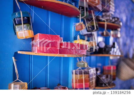 Colorful handmade soaps displayed on wooden shelves against a vibrant blue wall in a market setting, showcasing various shapes and sizes of soap bars Colorful handmade soaps displayed on wooden shelves against a vibrant blue wall in a market setting, showcasing various shapes and sizes of soap bars 139184256