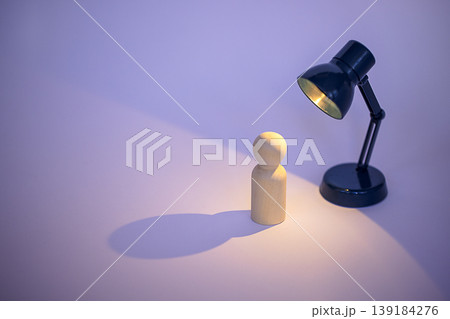 Wooden figurine stands under a black desk lamp casting a warm light and shadow on a smooth surface with a soft purple background Wooden figurine stands under a black desk lamp casting a warm light and shadow on a smooth surface with a soft purple background 139184276