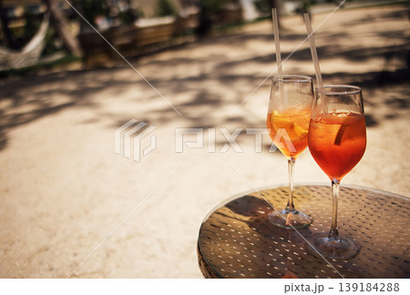 Two elegant glasses of aperol spritz cocktail with ice and straws placed on a round metal table in a sunny outdoor setting with blurred background Two elegant glasses of aperol spritz cocktail with ice and straws placed on a round metal table in a sunny outdoor setting with blurred background 139184288