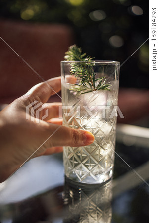 Hand holding a tall glass of refreshing cocktail with ice and a sprig of juniper, placed on a glass table in an outdoor setting with blurred background 139184293