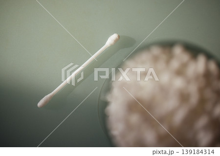 Cotton swab placed on a smooth surface next to a bowl filled with cotton balls, soft lighting enhances the clean and minimalistic aesthetic of the scene Cotton swab placed on a smooth surface next to a bowl filled with cotton balls, soft lighting enhances the clean and minimalistic aesthetic of the scene 139184314