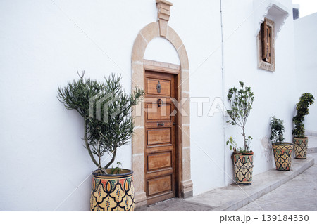 Wooden door framed by white wall, adorned with decorative planters, showcasing vibrant greenery and intricate patterns, creating a charming and inviting atmosphere in a serene outdoor setting Wooden door framed by white wall, adorned with decorative planters, showcasing vibrant greenery and intricate patterns, creating a charming and inviting atmosphere in a serene outdoor setting 139184330
