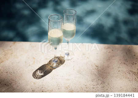 Two champagne flutes filled with sparkling beverage placed on a stone surface beside a swimming pool, with water reflections visible in the background Two champagne flutes filled with sparkling beverage placed on a stone surface beside a swimming pool, with water reflections visible in the background 139184441