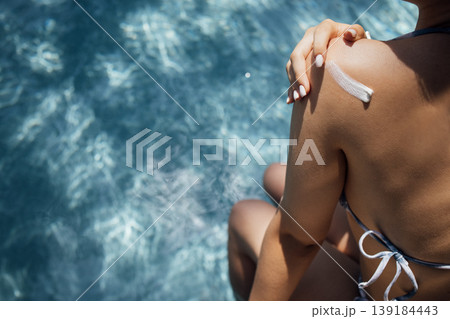 Female applying sunscreen on shoulder while sitting by the edge of a swimming pool with shimmering water reflecting sunlight in the background Female applying sunscreen on shoulder while sitting by the edge of a swimming pool with shimmering water reflecting sunlight in the background 139184443