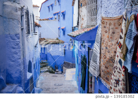 Narrow street in a vibrant blue city, adorned with traditional textiles hanging from walls, showcasing unique architecture and inviting atmosphere for exploration and cultural immersion 139185302