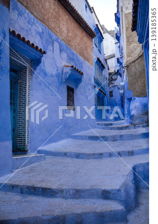 Blue-painted narrow alleyway in Chefchaouen, Morocco, featuring textured walls, winding steps, and traditional architecture under a clear sky 139185365