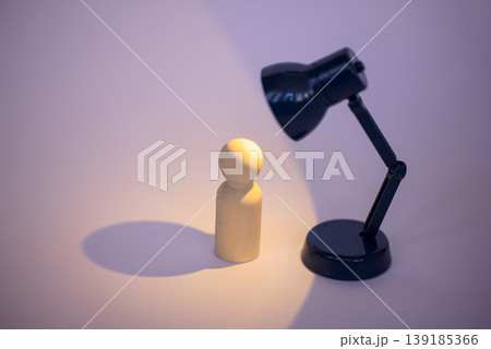 Wooden figurine illuminated by a black desk lamp casting a soft shadow on a light purple background, creating a minimalist and artistic composition 139185366