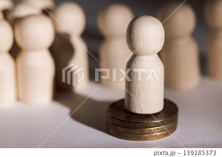 Wooden figurine standing on coins surrounded by blurred wooden figures, representing individuality and financial concepts in a minimalist setting Wooden figurine standing on coins surrounded by blurred wooden figures, representing individuality and financial concepts in a minimalist setting 139185373