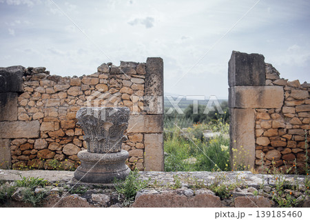 Ancient stone column base with intricate carvings in front of weathered stone ruins and green vegetation under a cloudy sky in a historical archaeological site Ancient stone column base with intricate carvings in front of weathered stone ruins and green vegetation under a cloudy sky in a historical archaeological site 139185460