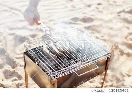 Close-up of a man hand holding a hand grill with fried sausages. Barbecue grill on the sandy beach. A man roasts sausages on an open fire. Close-up of a man hand holding a hand grill with fried sausages. Barbecue grill on the sandy beach. A man roasts sausages on an open fire. 139185491