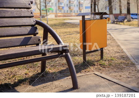 Yellow metal trash bin in a city park next to a brown wooden bench and a pedestrian path. Municipal metal trash container 139187201