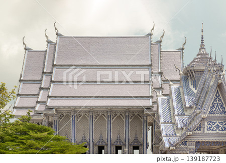 The sweeping, multi-tiered rooflines of Wat Pak Nam Khaem Nu Chanthaburi famed "Blue Temple" descend in rhythmic layers, their pale ceramic tiles shimmering softly beneath a hazy, overcast sky. Along 139187723