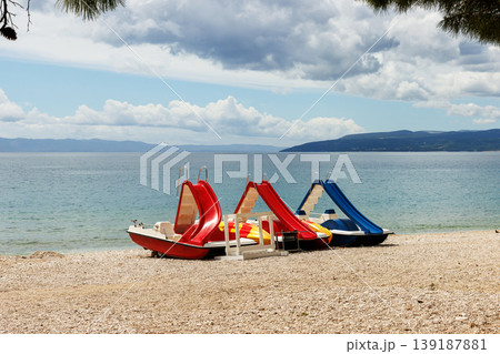 Colorful pedal boats on pebble beach by Adriatic sea in Croatia. Summer seaside leisure and travel scene with copy space. Colorful pedal boats on pebble beach by Adriatic sea in Croatia. Summer seaside leisure and travel scene with copy space. 139187881