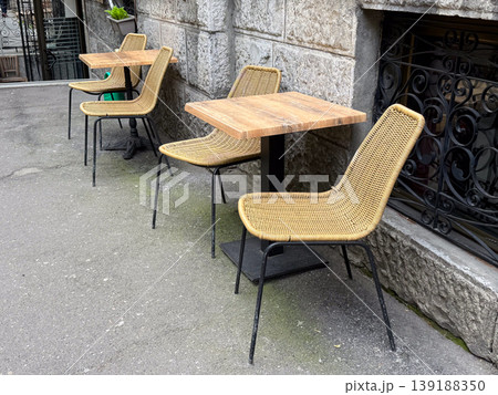 Outdoor cafe tables with woven chairs placed along stone wall on street. Urban lifestyle, relaxation, hospitality and dining environment concept. Outdoor cafe tables with woven chairs placed along stone wall on street. Urban lifestyle, relaxation, hospitality and dining environment concept. 139188350