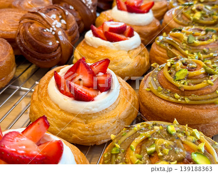 Danish pastries with strawberry cream and pistachio topping arranged closely on bakery rack. Sweet dessert selection, pastry variety, confectionery display and food styling concept. 139188363