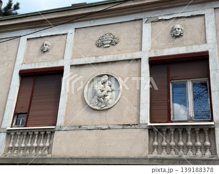 Old building facade with relief sculpture of woman and child surrounded by decorative elements. Architectural ornament, historical design, classical motif and urban heritage detail. 139188378