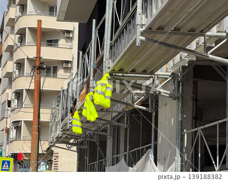 Construction scaffolding with fluorescent safety vests hanging near an unfinished building facade. Urban renovation, worker protection, building repair and construction site infrastructure. Construction scaffolding with fluorescent safety vests hanging near an unfinished building facade. Urban renovation, worker protection, building repair and construction site infrastructure. 139188382