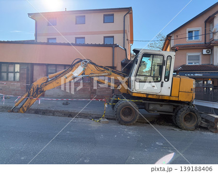 Wheeled excavator digging trench beside blocked sidewalk. Utility repair, street maintenance, construction zone, traffic restriction, infrastructure work, and residential inconvenience. Wheeled excavator digging trench beside blocked sidewalk. Utility repair, street maintenance, construction zone, traffic restriction, infrastructure work, and residential inconvenience. 139188406