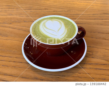 Matcha latte with heart foam art served in red cup on wooden table. Cafe beverage culture, barista craft, coffee alternative, and minimal composition. Matcha latte with heart foam art served in red cup on wooden table. Cafe beverage culture, barista craft, coffee alternative, and minimal composition. 139188442