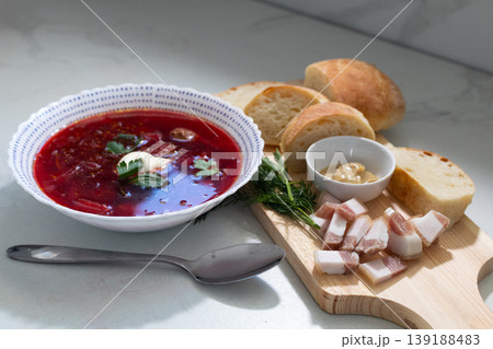 Bowl of traditional borscht soup topped with sour cream and fresh dill garnish. Served with rustic bread slices and smoked bacon on wooden 139188483