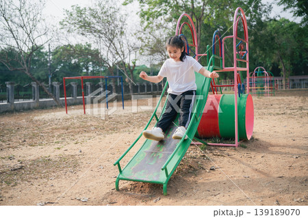 Happy Child Sliding Down a Colorful Playground Slide in an Outdoor Park Setting Under Clear Blue Skies Surrounded by Greenery and Play Equipment 139189070
