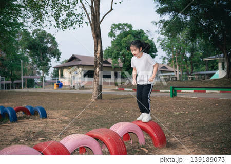 Energetic Child Playing on Colorful Tire Balance Course in Outdoor Playground Surrounded by Lush Greenery and School Facilities 139189073