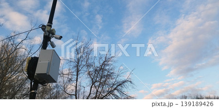 Security surveillance system mounted on black pole against a bright blue sky with wispy clouds. It features three CCTV cameras pointed in different directions and a large grey weatherproof utility box Security surveillance system mounted on black pole against a bright blue sky with wispy clouds. It features three CCTV cameras pointed in different directions and a large grey weatherproof utility box 139189426