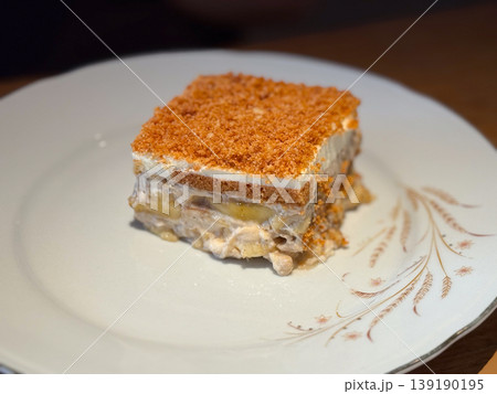 Piece of layered dessert with biscuit cream and fruit filling served on white plate in cafe interior. Sweet pastry, confectionery, snack serving, indulgence and culinary presentation. Piece of layered dessert with biscuit cream and fruit filling served on white plate in cafe interior. Sweet pastry, confectionery, snack serving, indulgence and culinary presentation. 139190195