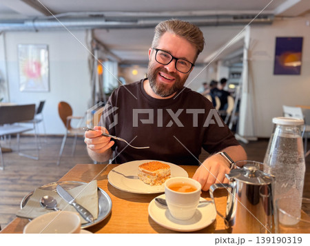 Smiling man with beard and glasses holding fork with dessert at cafe table with tea and tableware. Casual dining, enjoyment, social interaction, leisure moment and food experience. Smiling man with beard and glasses holding fork with dessert at cafe table with tea and tableware. Casual dining, enjoyment, social interaction, leisure moment and food experience. 139190319