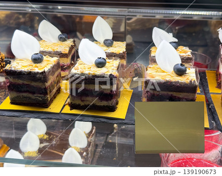 Layered cake squares with blueberry topping and white chocolate decoration. Dessert variety, pastry texture, confectionery detail, sweet assortment, and bakery presentation concept. 139190673