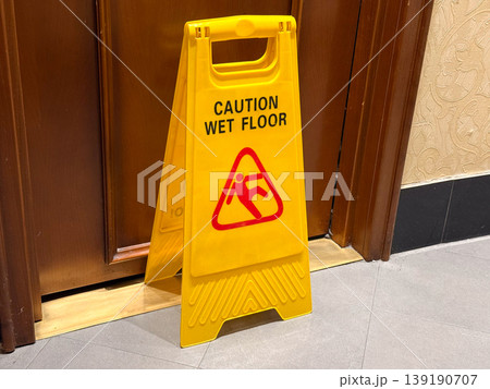 Yellow caution wet floor sign placed on indoor surface. Safety warning, hazard prevention, public awareness, cleaning process, and risk management in interior space. Yellow caution wet floor sign placed on indoor surface. Safety warning, hazard prevention, public awareness, cleaning process, and risk management in interior space. 139190707