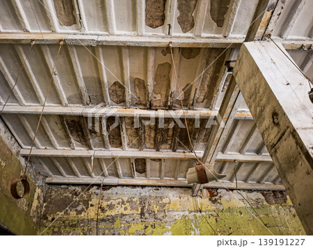 Derelict interior shows a ceiling with peeling paint and exposed concrete in an abandoned warehouse. Hanging metal fixtures add to the decayed look of the structure. Derelict interior shows a ceiling with peeling paint and exposed concrete in an abandoned warehouse. Hanging metal fixtures add to the decayed look of the structure. 139191227