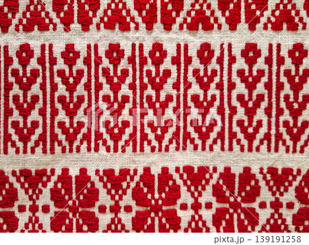 Close up view of white linen fabric featuring bright red geometric embroidery. The design shows classic patterns commonly found in Slavic or Eastern European art. Close up view of white linen fabric featuring bright red geometric embroidery. The design shows classic patterns commonly found in Slavic or Eastern European art. 139191258