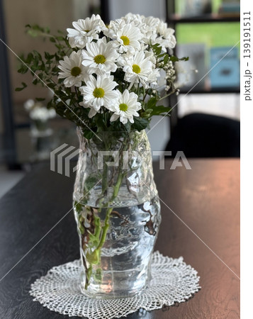 Glass vase with chrysanthemum bouquet placed on wooden table with bookshelf background in cafe interior. Floral decoration, home styling, botanical composition, natural freshness and cozy atmosphere. 139191511
